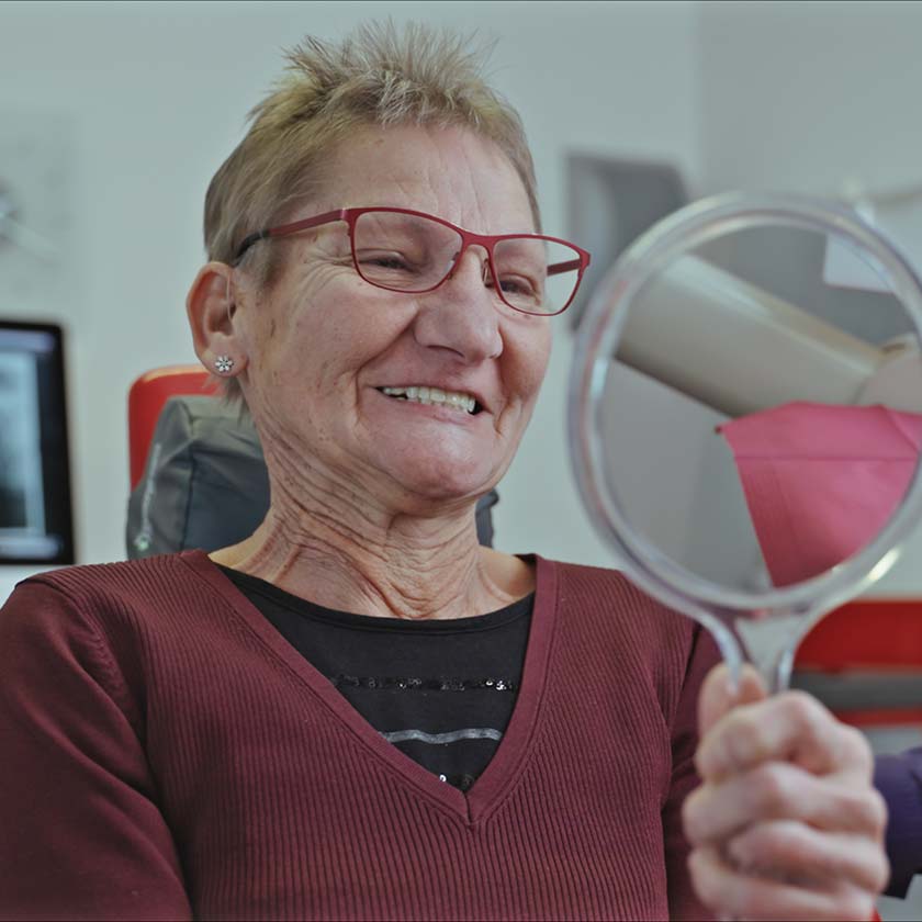 Patientin hält Handspiegel in der Hand und betrachtet im Behandlungsraum der 5-Sterne Praxis Dr. Gal nach der Feste Zähne an einem Tag Behandlung ihre Zähne
