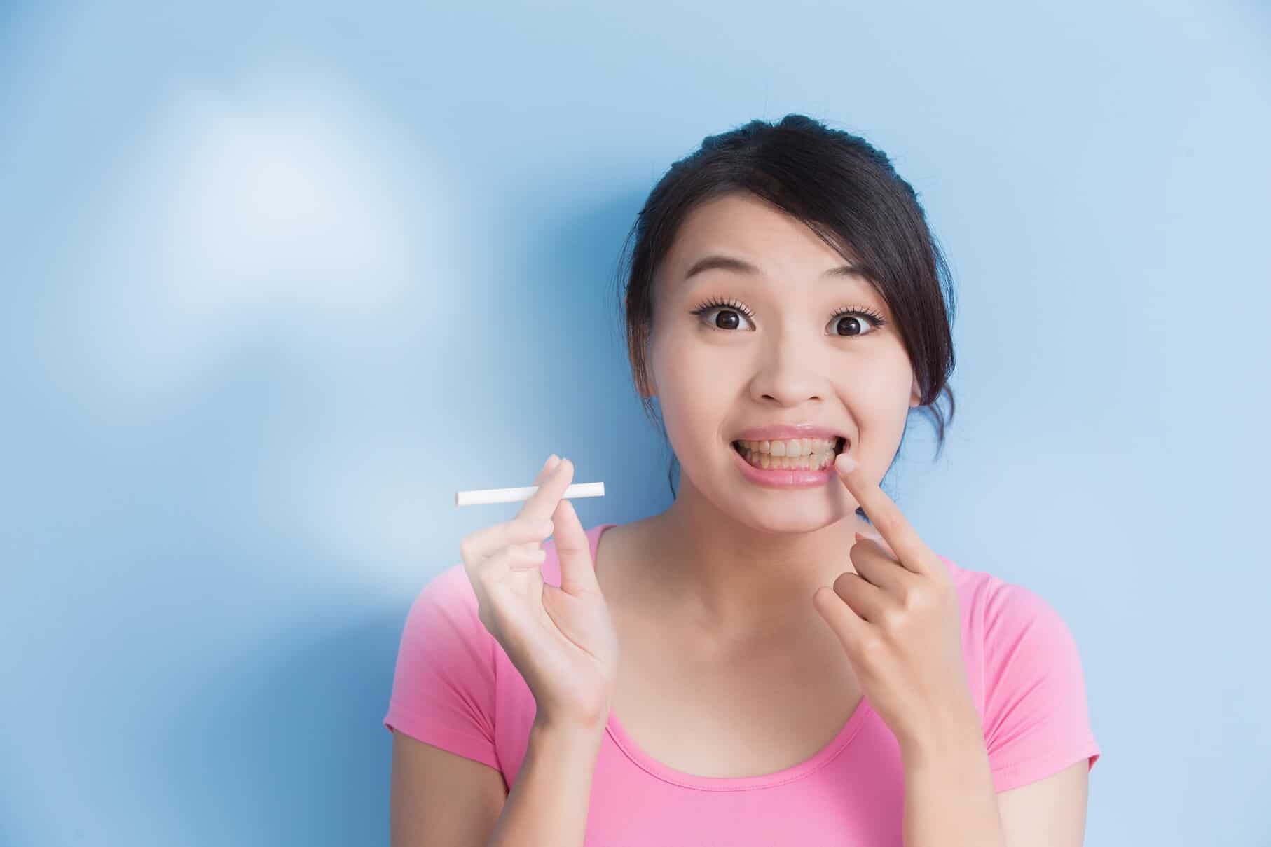 Junge asiatische Frau macht sich zu spät Gedanken über Zahngesundheit, sie hält in der rechten Hand eine Zigarette und zeigt mit der linken Hand auf ihre gelben Zähne