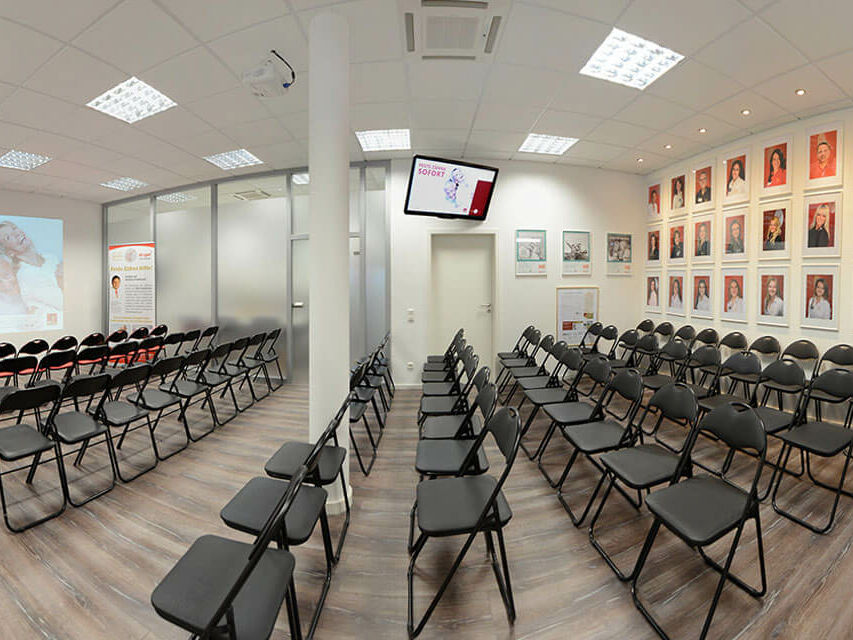 Raum mit Stuhlreihen ausgestattet, vorne eine Leinwand, hinten an der Wand hängen die Bilder der Mitarbeiter der Zahnarztpraxis Dr. Gal